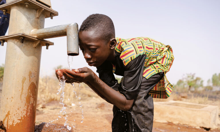 Thirsty young black African boy leans forward to drink fresh and clean water with his bare hands from a tap; water scarcity concept